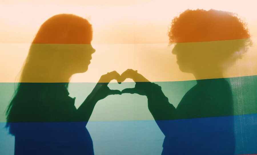 Two girls making heart shape using their hands, behind LGBT flag