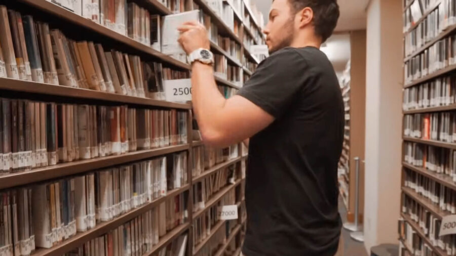 A man in a black t-shirt is organizing or retrieving a book or a DVD from a shelf in a library or archive. The environment appears quiet and orderly, suitable for studying or research.