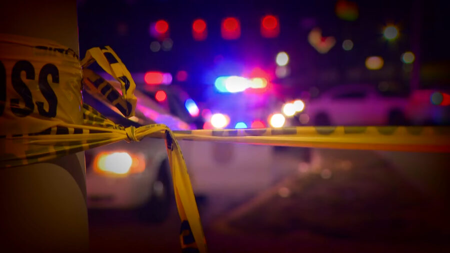 Close-up of yellow crime scene tape tied around a pole, with blurred police car lights flashing in the background during the night.