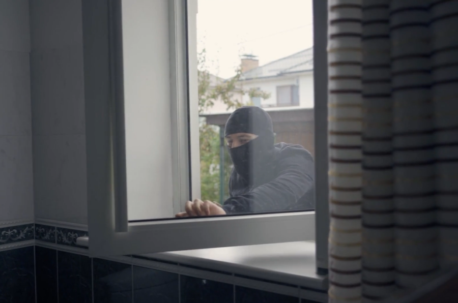 A masked individual wearing dark clothing is seen attempting to break into a house through an open window. The scene is viewed from inside the house, highlighting the person's hands gripping the window frame.