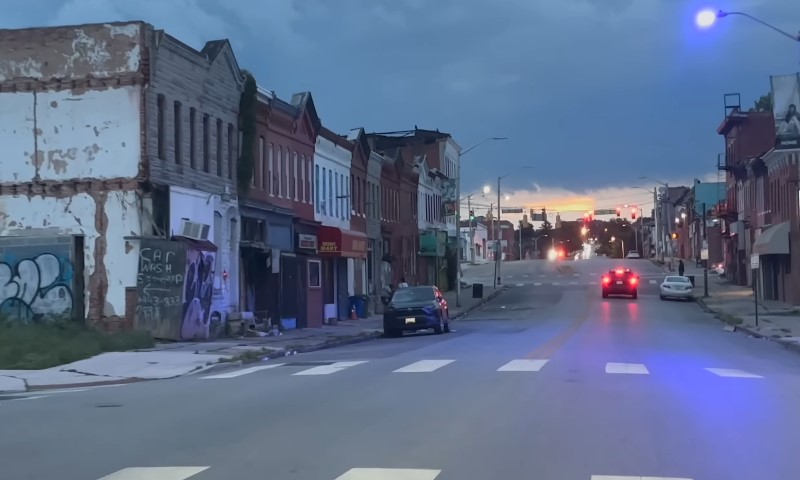Baltimore street scene at dusk with aging rowhouses and car taillights glowing red