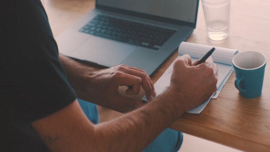 Man writing on paper with laptop in the background