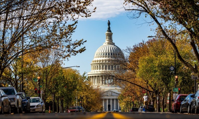 U.S. Capitol dome framed by autumn trees along a street, with parked cars and a pedestrian