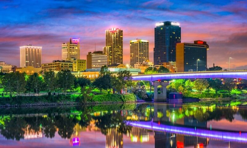 Twilight cityscape of Little Rock, Arkansas, with illuminated skyscrapers and a lit bridge reflecting vibrantly in the calm river below