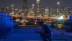 A police officer crouches on a dimly lit sidewalk at night, examining the crime scene. City skyline glows in the background