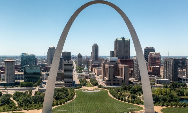 Aerial view of the St. Louis Gateway Arch in a park setting, with a clear blue sky, surrounded by a city skyline of tall buildings