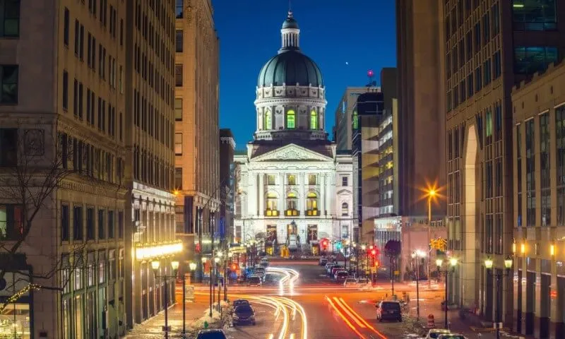 A majestic domed building, brightly lit, stands at the end of a bustling city street in Indianapolis.
