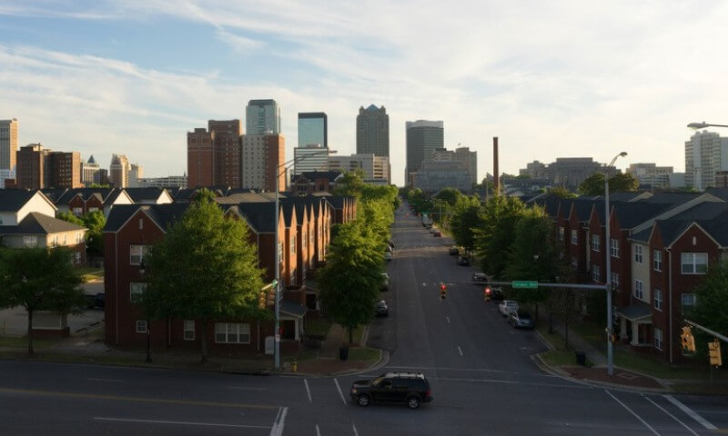 Urban street scene with red brick townhouses lining both sides of the road, lush green trees, and a distant skyline