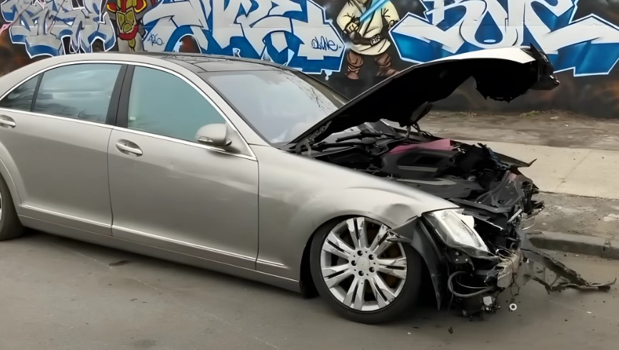 A silver Mercedes-Benz sedan, significantly damaged in an accident, sits on the street