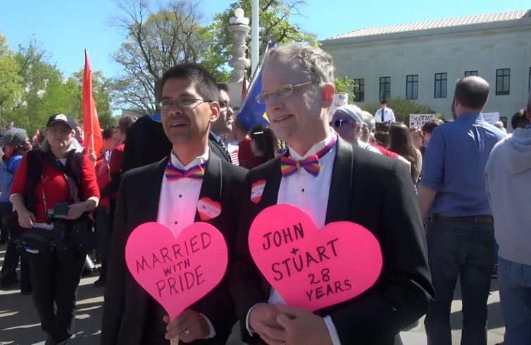 Two men, celebrating 28 years of marriage, hold signs that read "Married with Pride" and "John + Stuart 28 Years