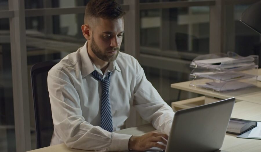 A concentrated worker hunches over a laptop, surrounded by paperwork