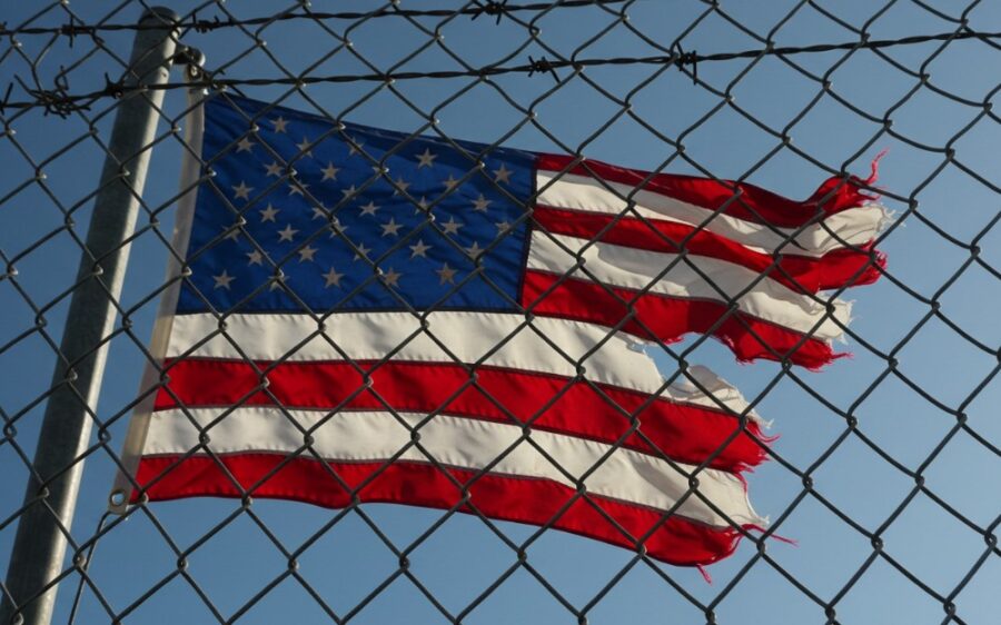 The American flag, partially torn, billows slightly behind a chain-link fence