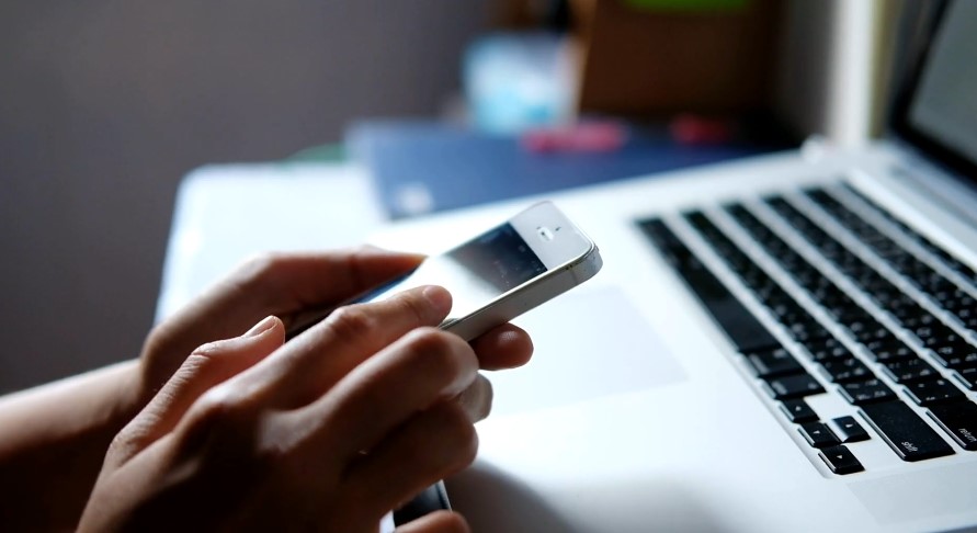 Close-up view of hands using a smartphone while a laptop is visible in the background