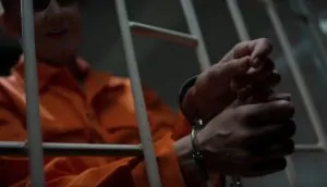 A person in an orange jumpsuit, handcuffed, sits within a metal cage