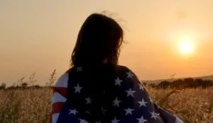 A young woman stands in a golden field, the American flag draped around her shoulders, as the sun sets