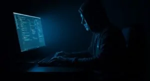 A hacker hunches over a computer screen, illuminated by the glow of lines of code