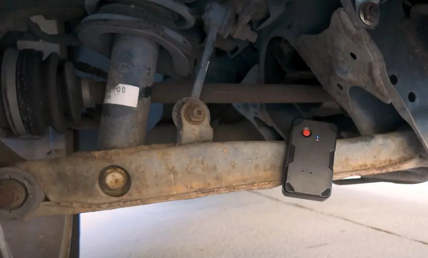 A small, black tracking device rests beneath a rusty vehicle's chassis