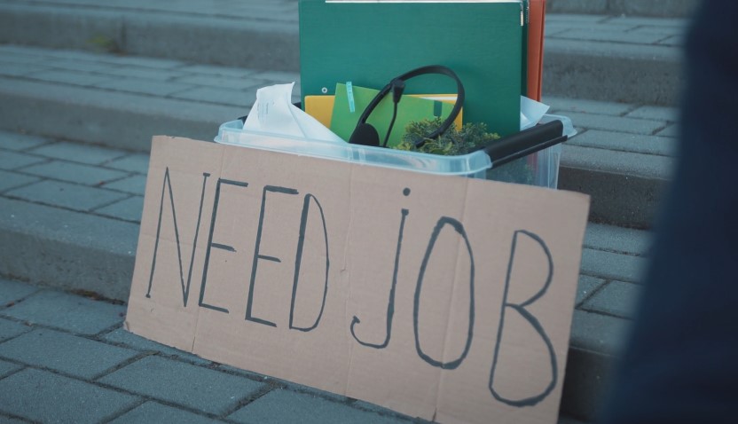 A cardboard sign, pleading "NEED JOB," sits atop a collection of personal items