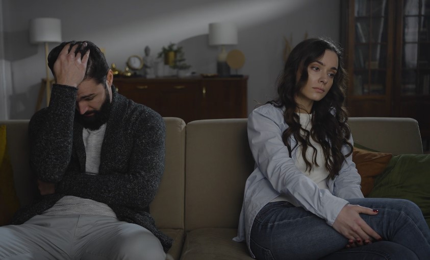 A couple sits apart on a couch, their expressions conveying a sense of distance and quiet distress