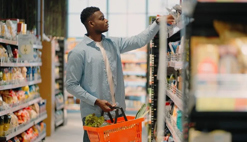 A man carefully selects items from the grocery store shelves, his basket filled with fresh produce