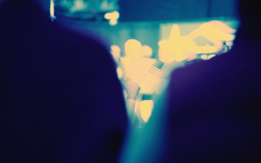 People hold signs and flags at a political rally, seen through a crowd in low light