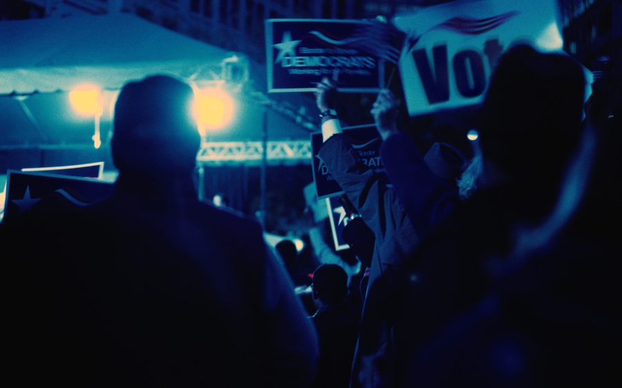 People hold signs and banners during a political rally at night under bright lights