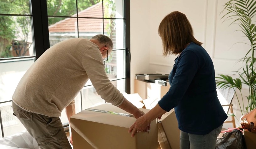 A couple works together, carefully lifting a large cardboard box
