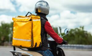 A food delivery driver on a scooter wears a helmet and yellow backpack, reflecting changes under Boston’s new delivery regulations