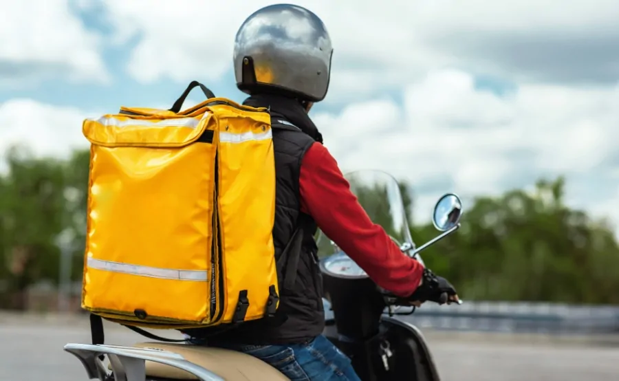 A food delivery driver on a scooter wears a helmet and yellow backpack, reflecting changes under Boston’s new delivery regulations