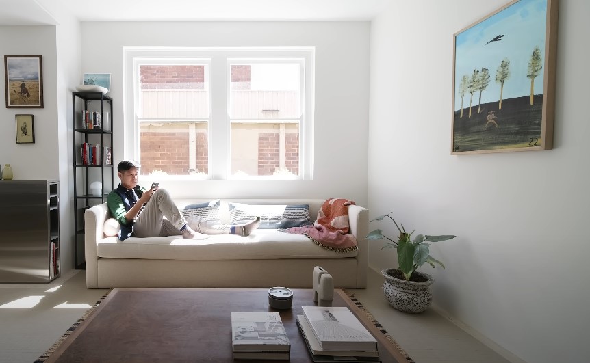A man relaxes on a light beige couch, engrossed in his phone, in a bright, airy living room