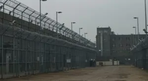 A vast, gray expanse of a correctional facility, enclosed by razor wire and concrete walls