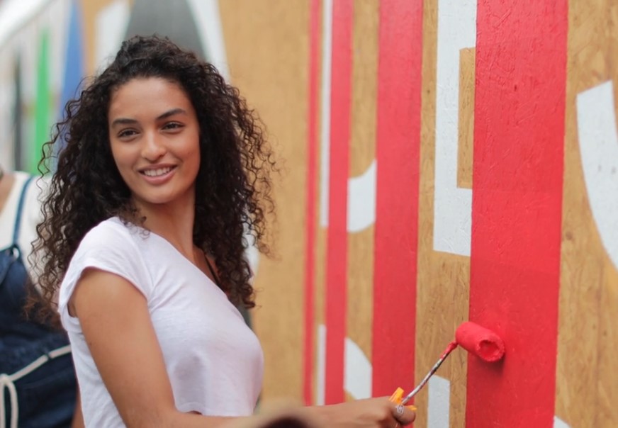 A woman applies vibrant red paint to a large-scale mural, adding a pop of color to the urban landscape