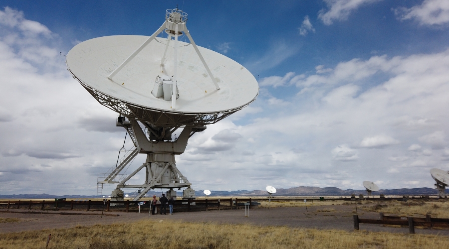 A large satellite dish stands in an open field with several smaller dishes visible in the distance under a partly cloudy sky