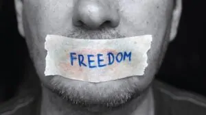 A close-up of a man with tape over his mouth labeled “FREEDOM,” symbolizing restrictions on free speech