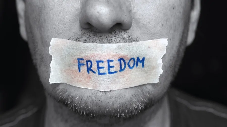 A close-up of a man with tape over his mouth labeled “FREEDOM,” symbolizing restrictions on free speech