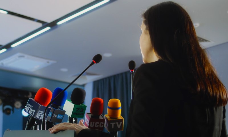 A woman stands behind a podium with multiple microphones, addressing an audience
