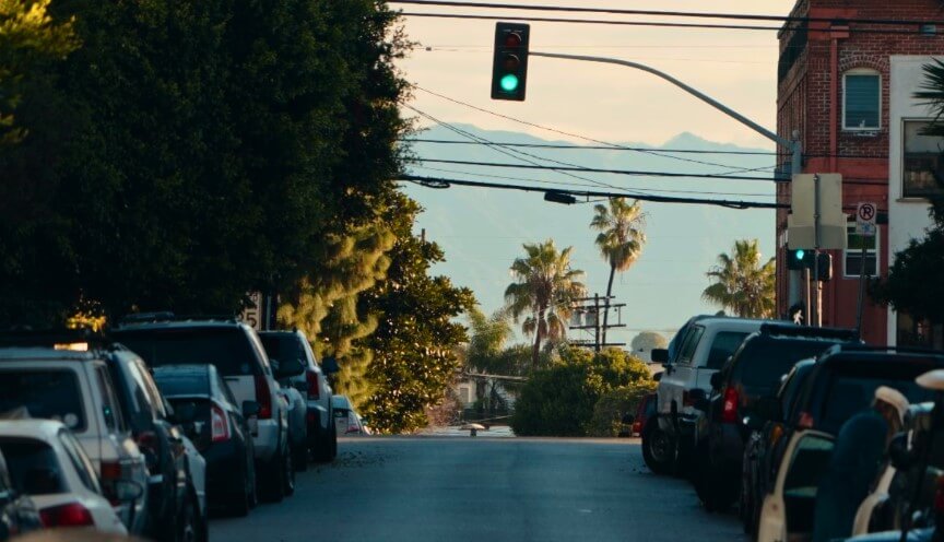 Green light, palm trees, and a street lined with parked cars under a hazy sky