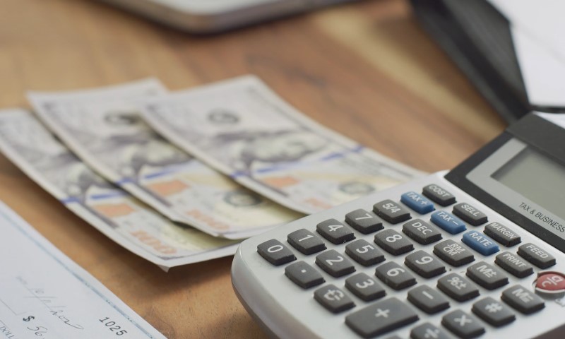 Close-up of calculator keys next to stacked US hundred-dollar bills and a partially visible check on a wooden surface