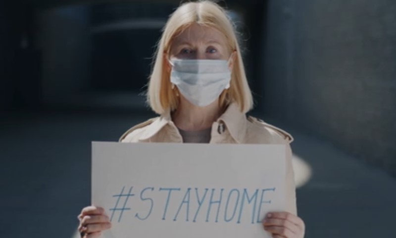 A woman in a face mask holds a sign reading "Stay Home"