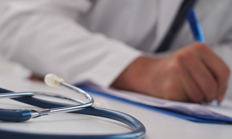 A doctor in a white coat writes on paper while stethoscope is on the table