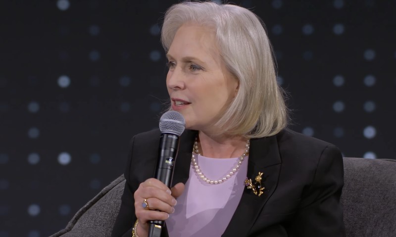Senator Gillbrand in a black shirt and pearls holds a microphone