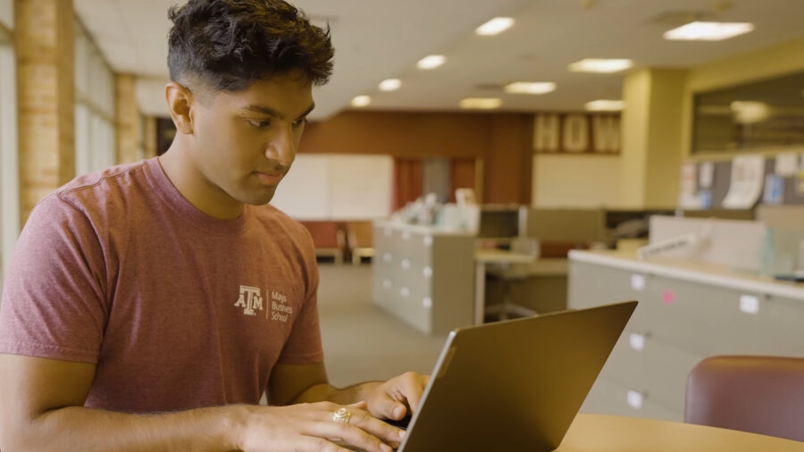 A man in a maroon shirt sits at a table, working on a laptop