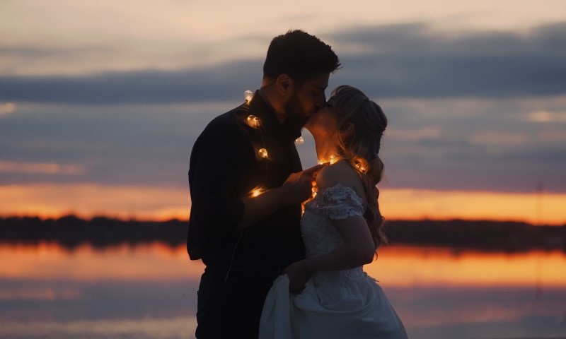 A bride and groom share a kiss at sunset, surrounded by twinkling lights creating a romantic atmosphere