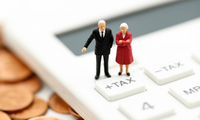 Miniature elderly couple standing on a calculator's "+TAX" button near scattered coins, symbolizing retirement financial planning