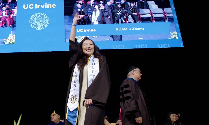 A woman in a graduation gown waves to the audience, celebrating her achievement during the ceremony
