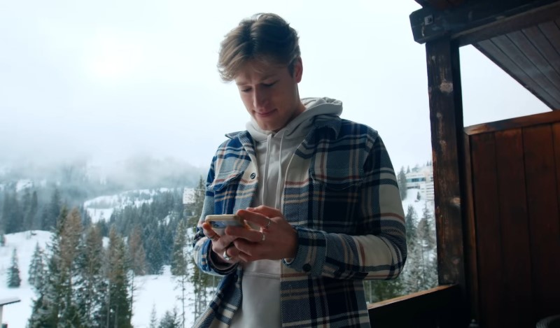 A man stands on a balcony, gazing at his phone with a thoughtful expression