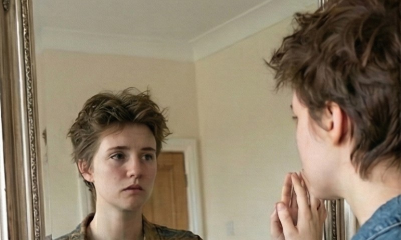 A person with short hair gazes thoughtfully at themselves in a mirror