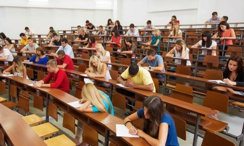Students seated in a large lecture hall take an exam, focused on writing answers