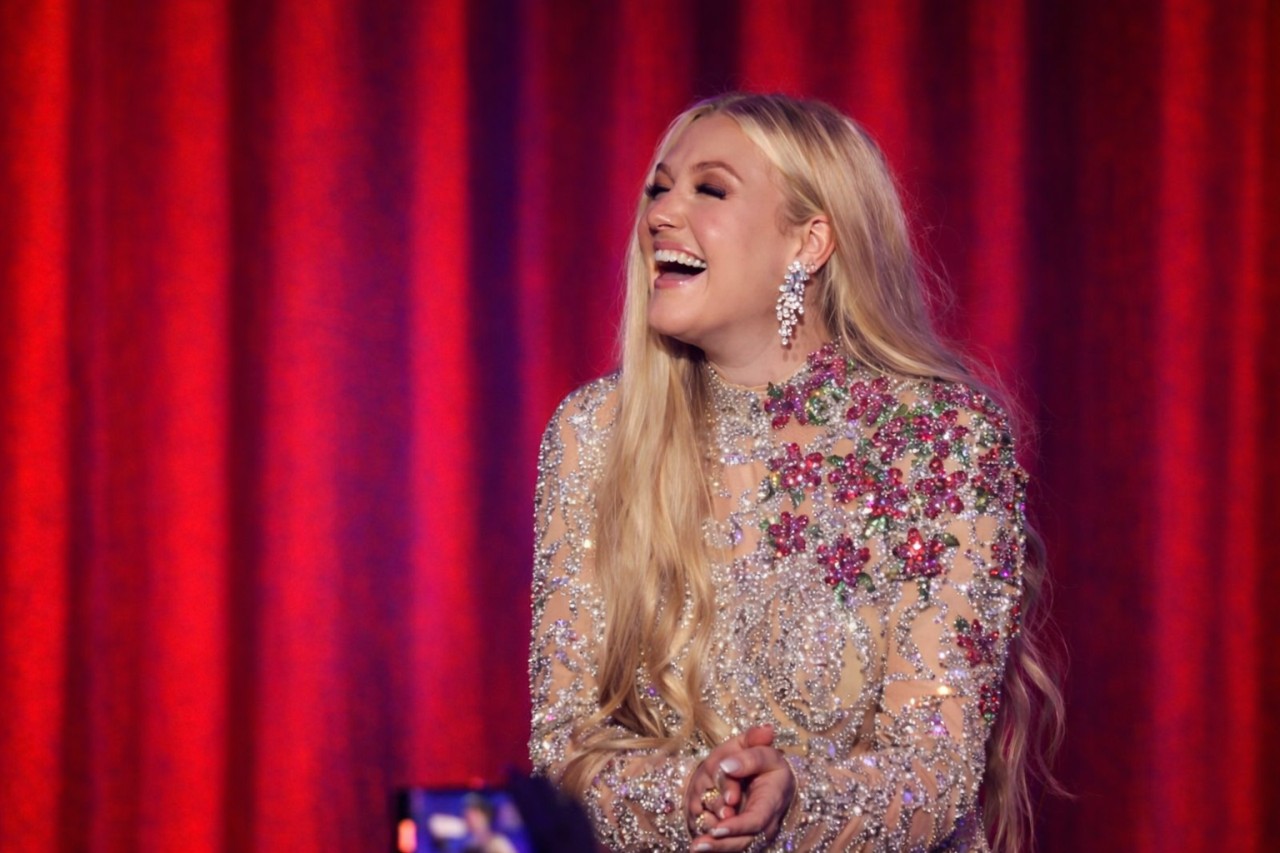 Erika Kirk with long blonde hair laughs joyfully, wearing a sparkling gown adorned with colorful floral designs