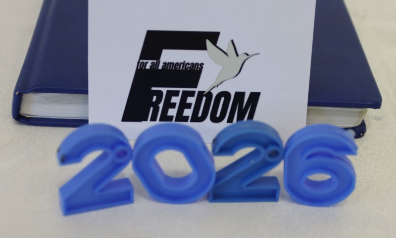 Blue "2026" stands before a card with "Freedom for all Americans" text and a bird graphic. A closed blue book serves as the backdrop.